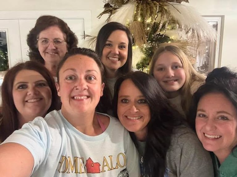 Group of seven smiling women posing together indoors with holiday decorations visible in the background