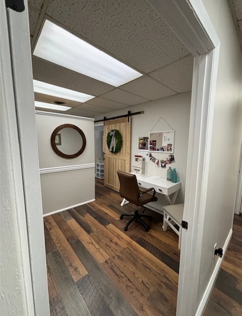 Modern home office interior with wooden flooring, white desk, black office chair, and wall decorations including a round mirror and wreath