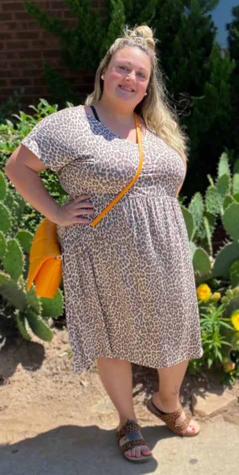 Woman wearing red plaid shirt and blue jeans standing against brick wall, smiling at camera with sunglasses on head