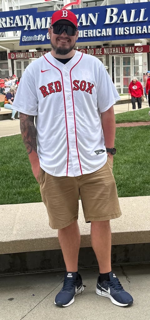 Man wearing Boston Red Sox baseball jersey and cap standing at a stadium with signage in background