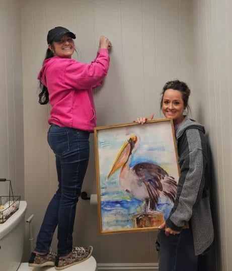 Two people hanging a framed watercolor painting of an eagle on a white wall indoors