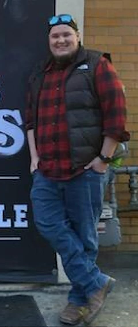 Person wearing red plaid shirt, black vest, and blue jeans standing against a brick wall, smiling at camera with sunglasses on head