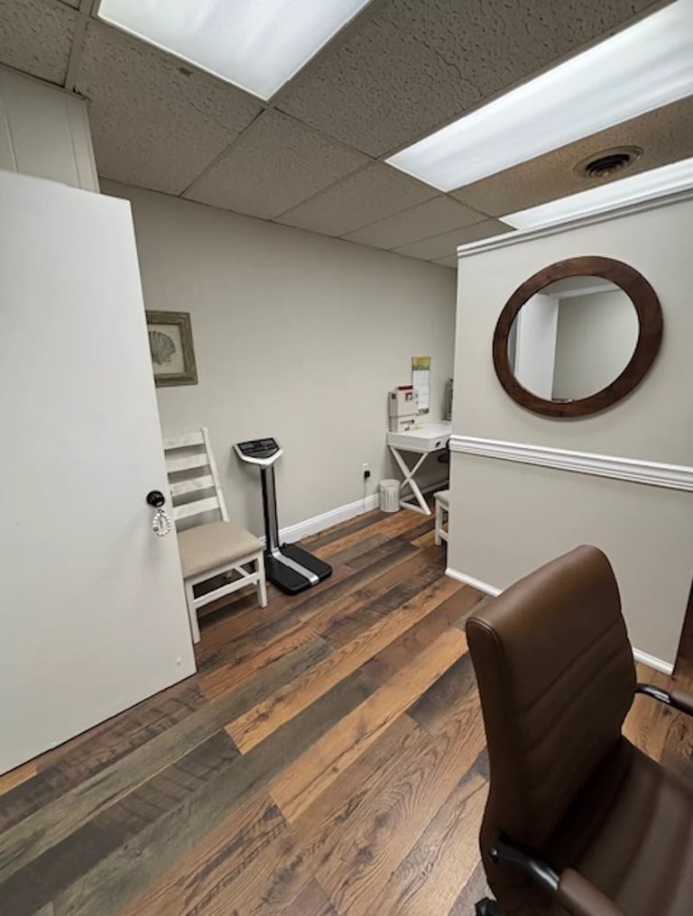 Interior of a small medical or office space with wooden flooring, white walls, skylight ceiling, exercise equipment, and brown circular mirror