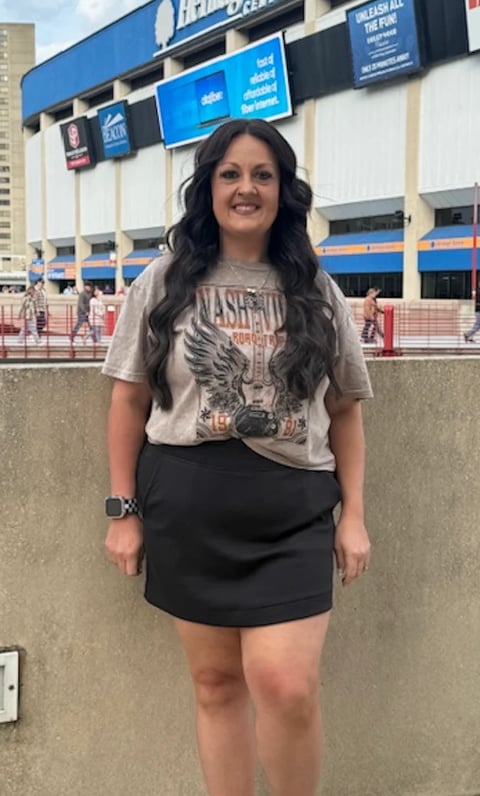 Woman smiling at camera wearing graphic t-shirt and black skirt, standing in front of sports arena